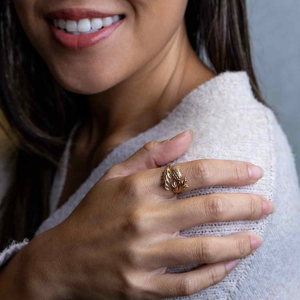Bronze ring with thistle flower on the index finger of a right hand leaning on a left shoulder of a smiling woman of color with loose, sleek brown hair.