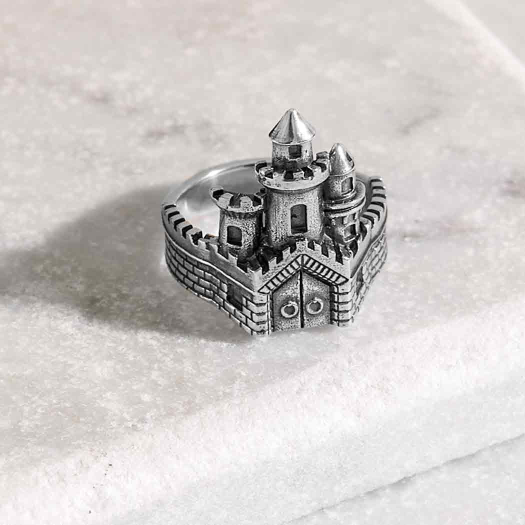 Photograph of a silver castle ring including three turrets and battlements on a marble base. The castle ring wall goes around the finger.